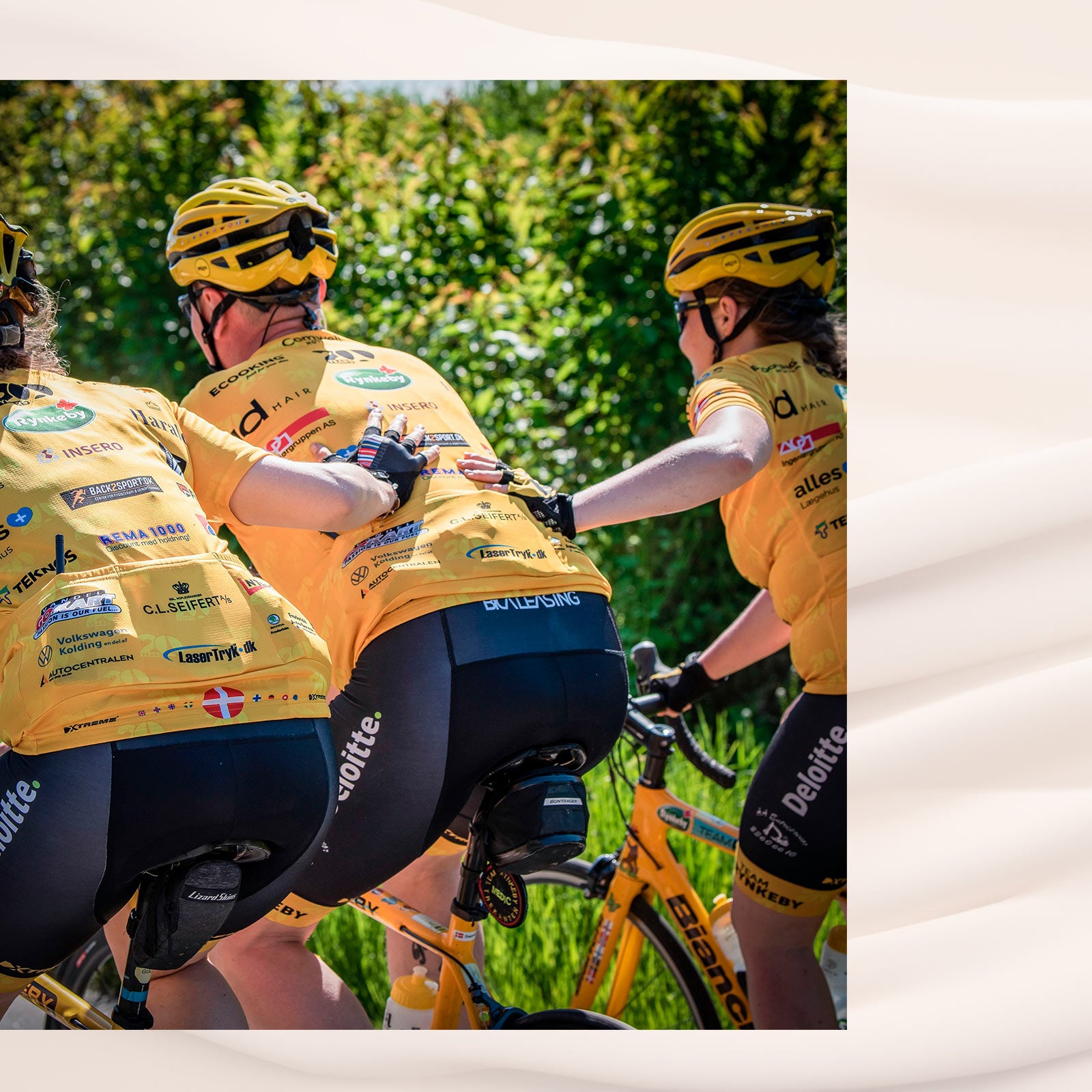 Tre cykelryttere i gule trøjer med sponsorer kører tæt sammen og støtter hinanden.