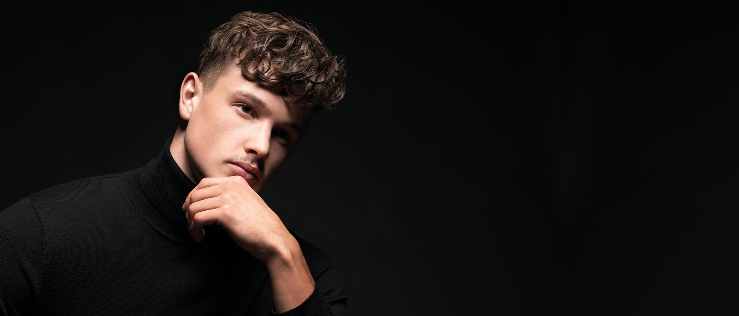 Mand med kort, bølget hår og mørk rullekrave mod sort baggrund.