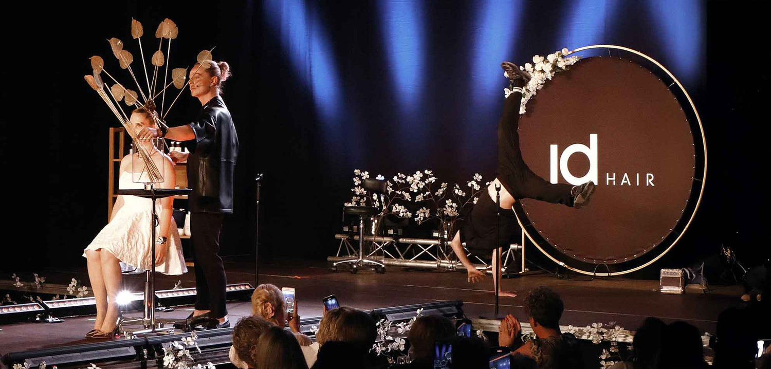 Stylist arbejder på model med kunstnerisk hårudsmykning på IdHAIR-scene, mens performer laver håndstand ved stort IdHAIR-logo