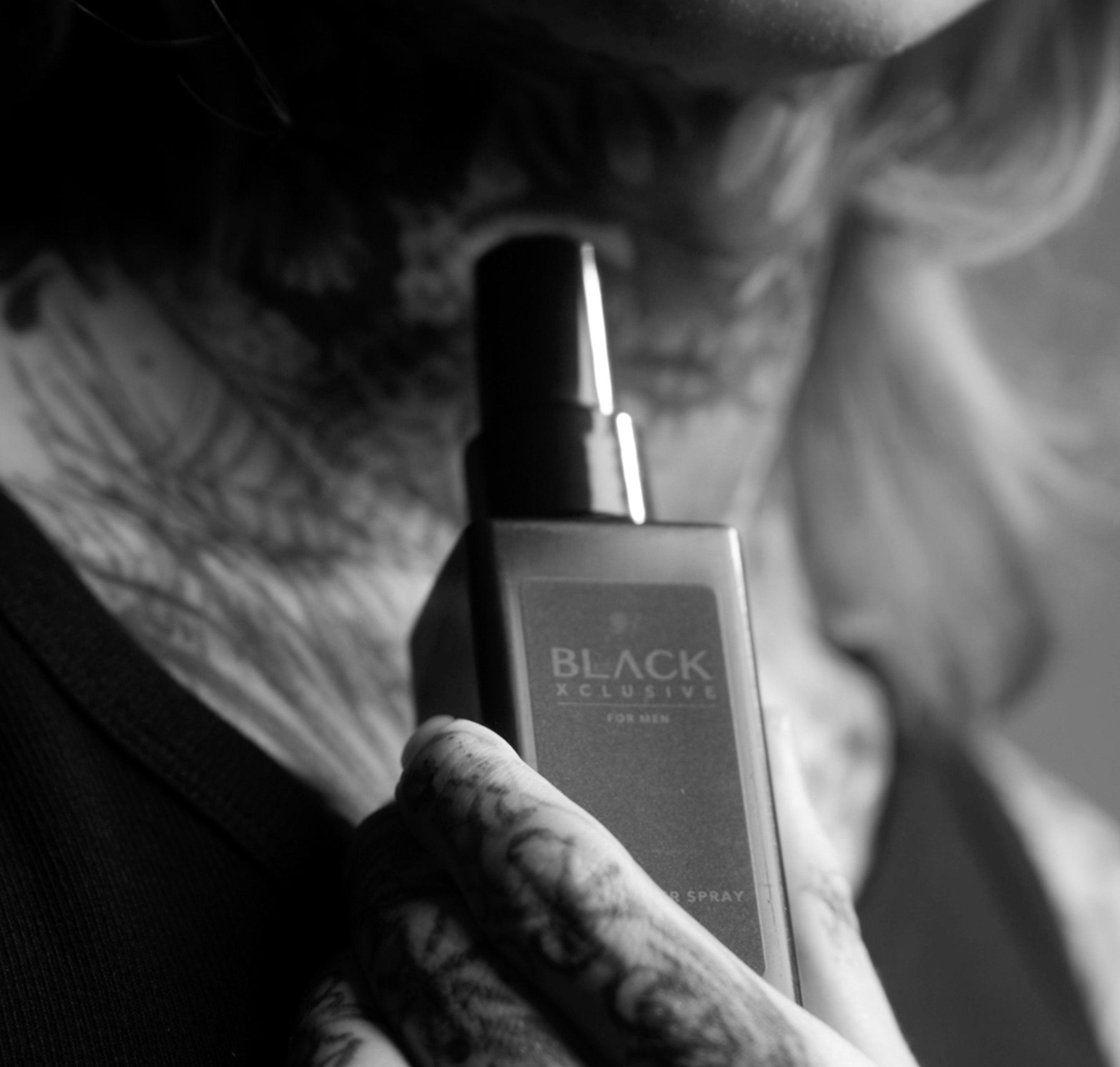 Sort flaske med Black Xclusive Saltwater Spray holdt i en tatoveret hånd. Stilfuldt sort-hvidt billede med rå og edgy æstetik