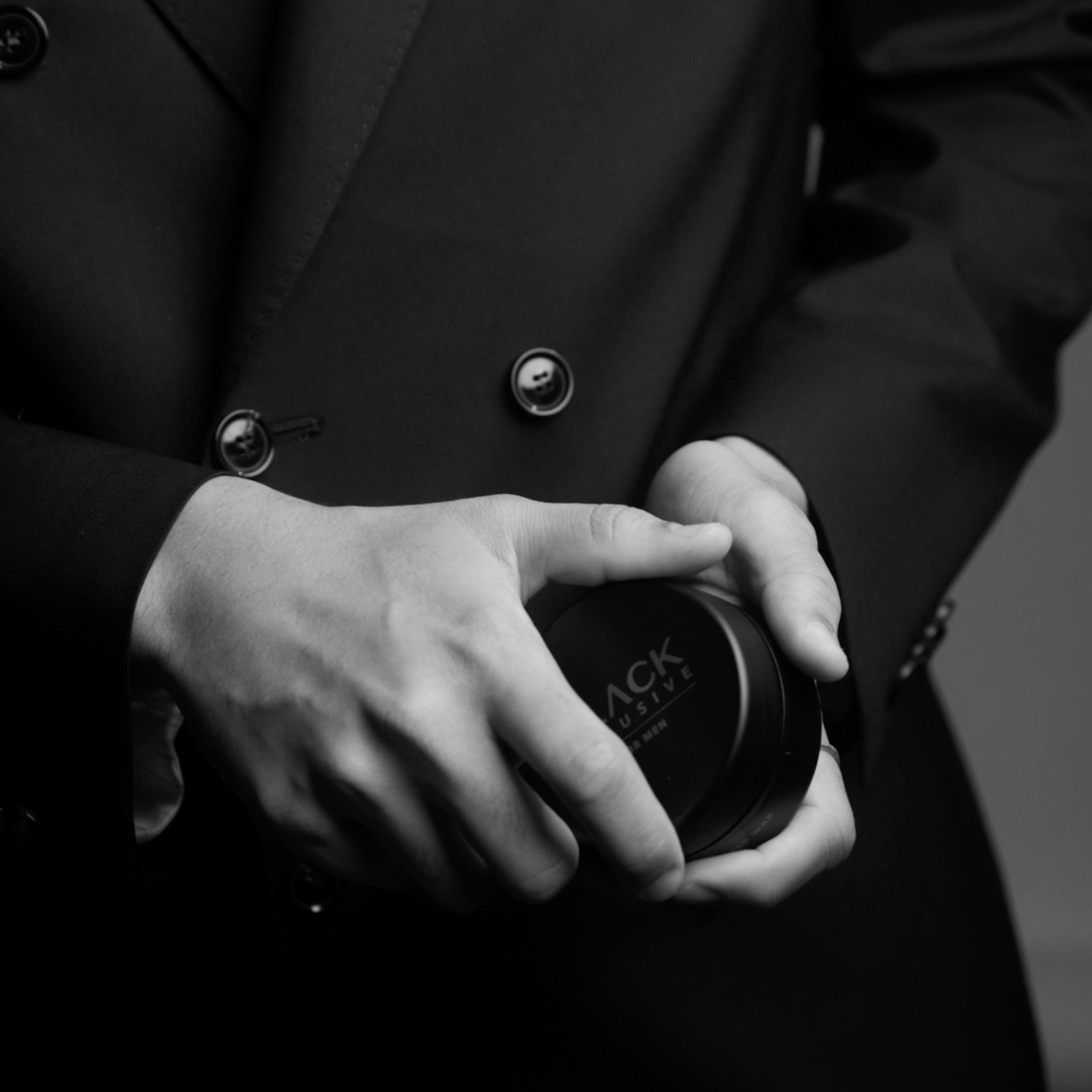 Black Xclusive Wax fra IdHAIR holdt i hænderne på en person iført en stilfuld sort blazer. Elegant sort-hvidt billede med et maskulint og eksklusivt udtryk.