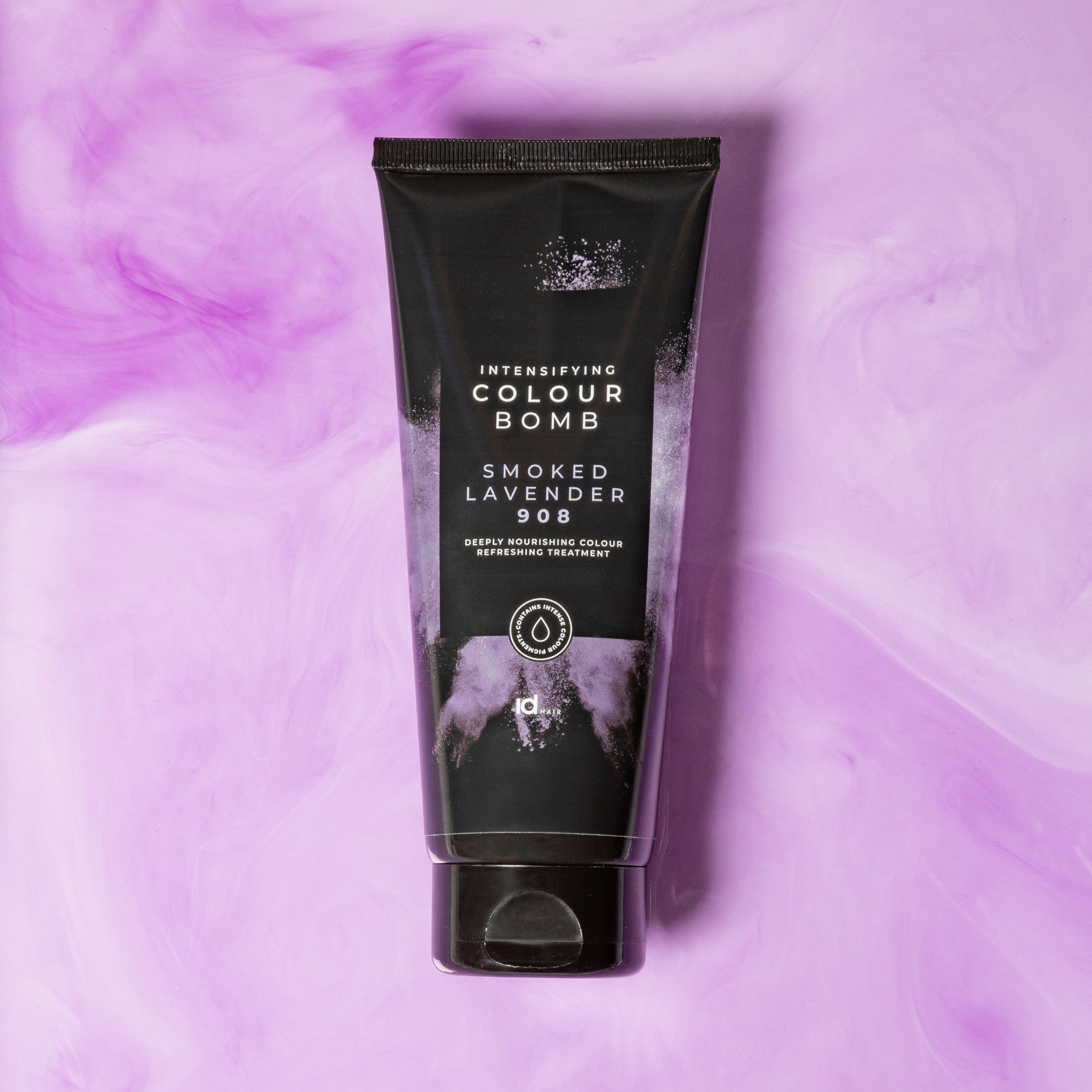 Colour Bomb Smoked Lavender 908 farvekur fra IdHAIR, placeret på en swirlende lavendelfarvet baggrund. Stilfuld opstilling, der fremhæver produktets dybe, lilla toner.