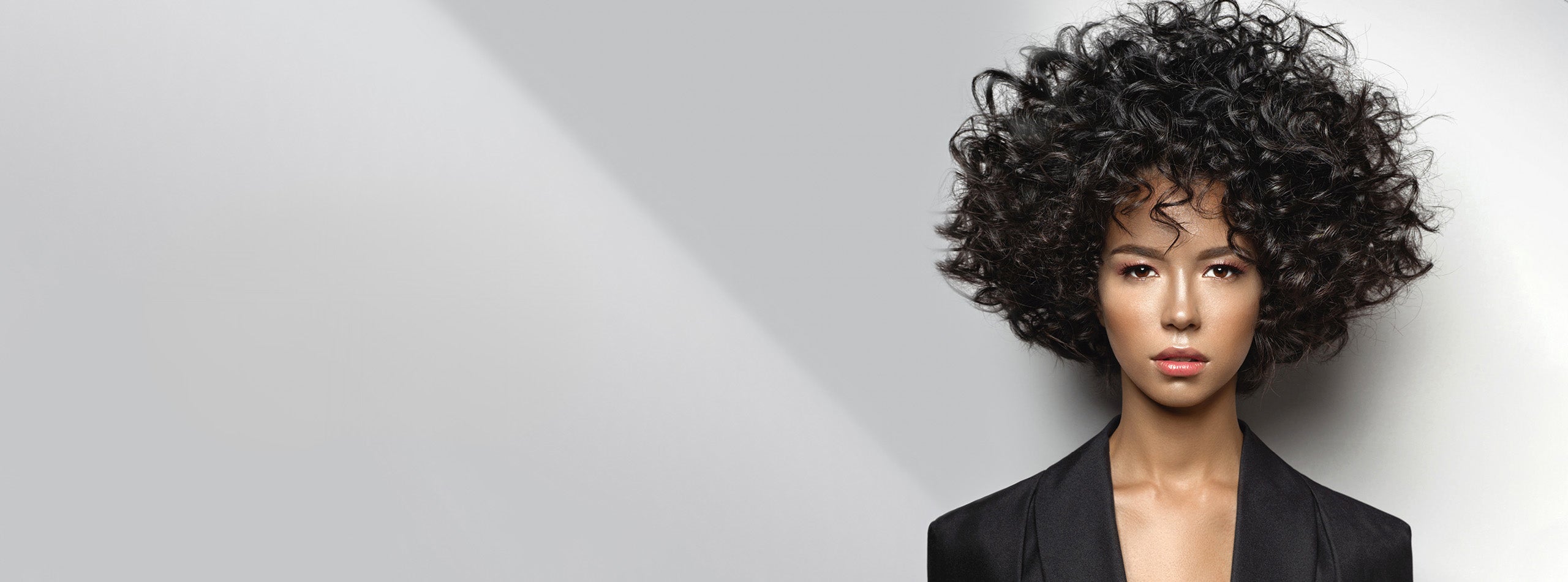 Kvinde med kort, mørkt og fyldigt krøllet hår mod grå baggrund.